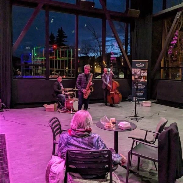 The Edward residents enjoying a performance from a trio of musicians. The musicians are playing a drum kit, saxophone, and upright bass.