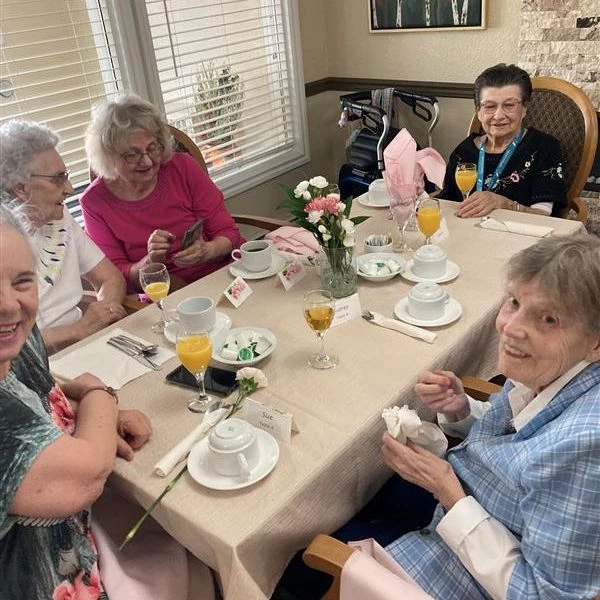 Multiple senior women enjoying some tea and drinks together. Two of the women are looking at a phone, while the others are looking at the camera and smiling.