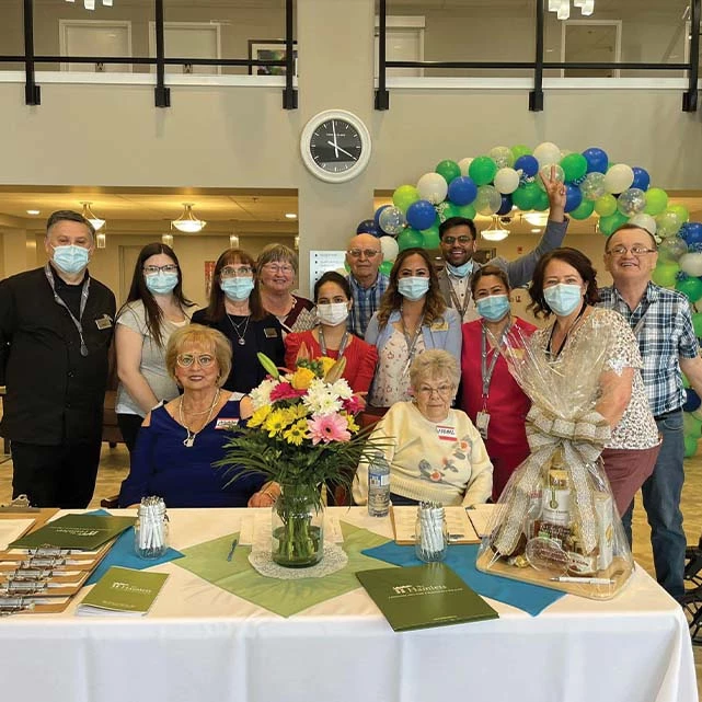 A table with two seniors seated at it. Behind the seniors are a bunch of The Hamlets at Red Deer staff posing behind them. Further back, there is a large balloon arch.