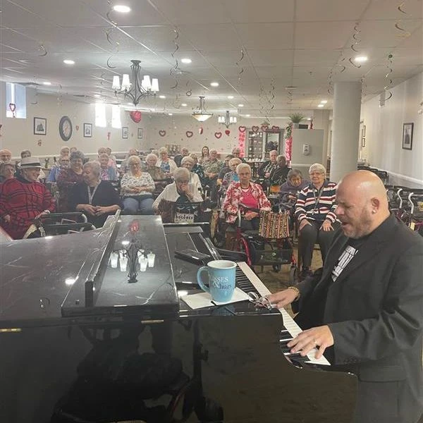 A man playing the piano and singing while seniors are watching the performance. There are Valentine's Day decorations hanging from the roof as well.